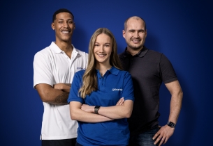 Drei lächelnde Personen in Arbeitskleidung vor blauem Hintergrund, stehen zusammen und strahlen Teamgeist aus.