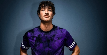 Junger Mann in lila T-Shirt posiert vor einer blauen Wand, lächelt selbstbewusst.